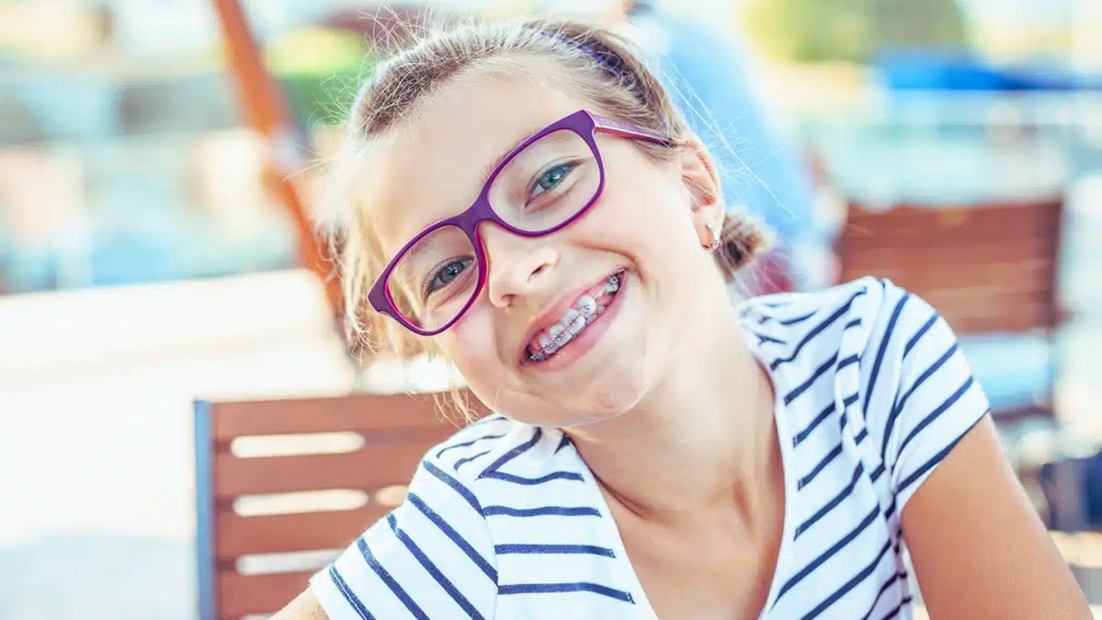 A smiling young girl wearing glasses and braces sits at a table, exuding happiness and a cheerful demeanor.