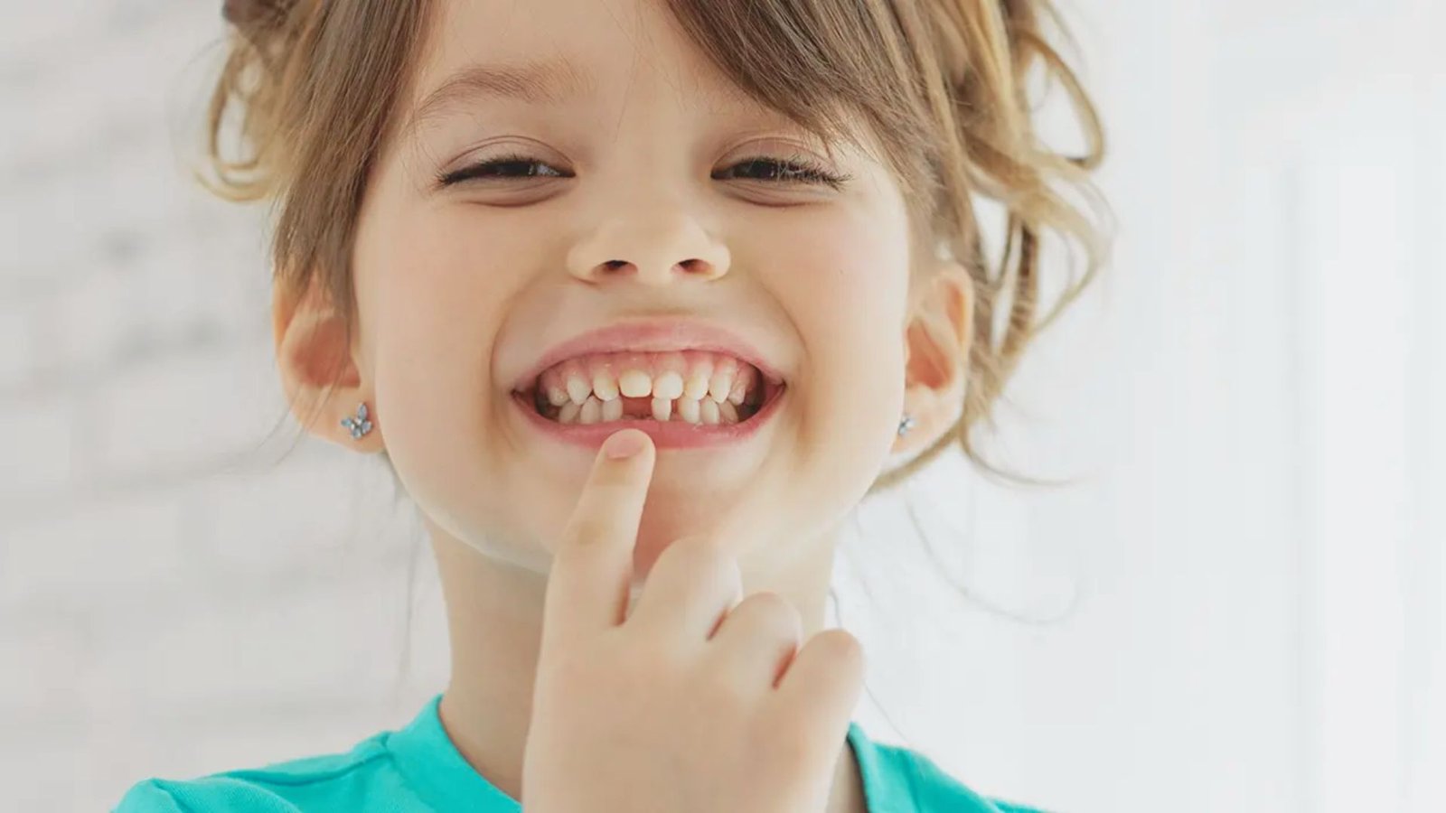 A cheerful young girl smiles while placing her finger to her lips, indicating a playful gesture.