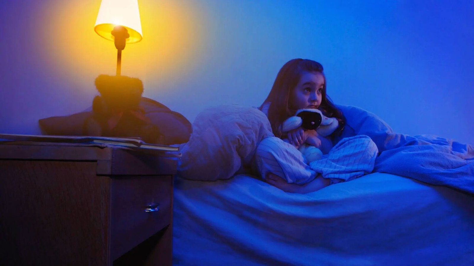 A girl sitting in bed with a lamp on and a phone in her hand, contemplating fears of the dark.