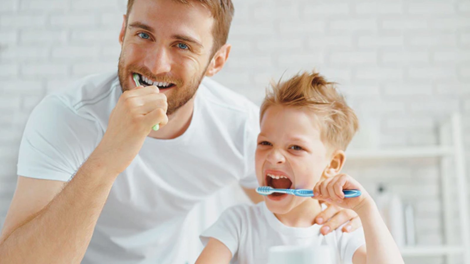 Father and young child brushing their teeth together in a bright bathroom, both smiling while holding toothbrushes.