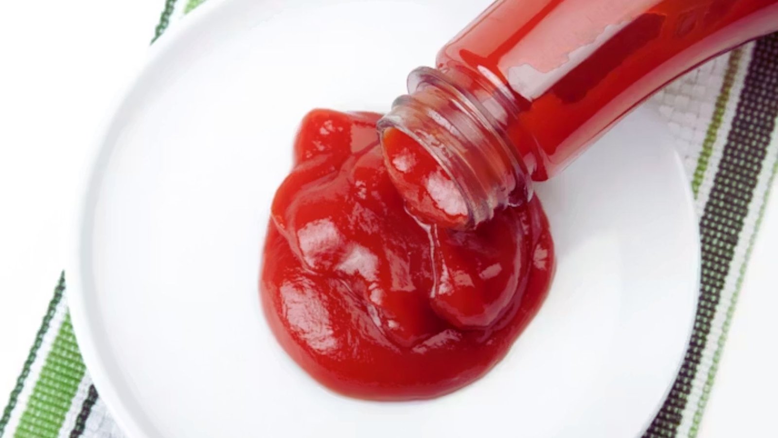 A close-up of ketchup being poured from a bottle onto a white plate, creating a vibrant red swirl.