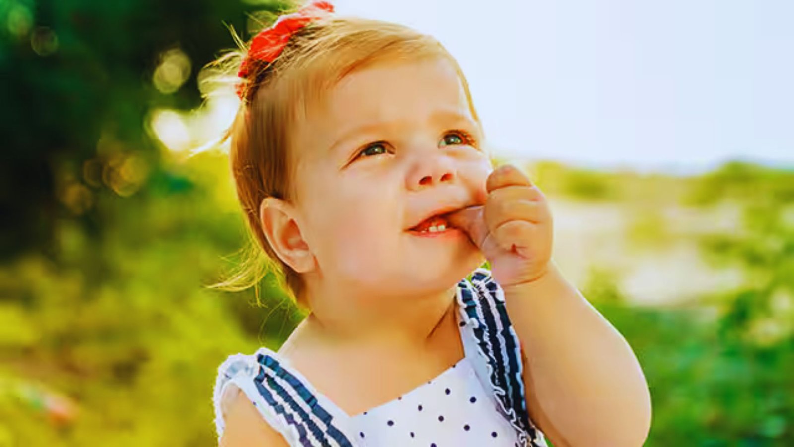 A baby girl stands in the grass, playfully biting her finger with a curious expression.