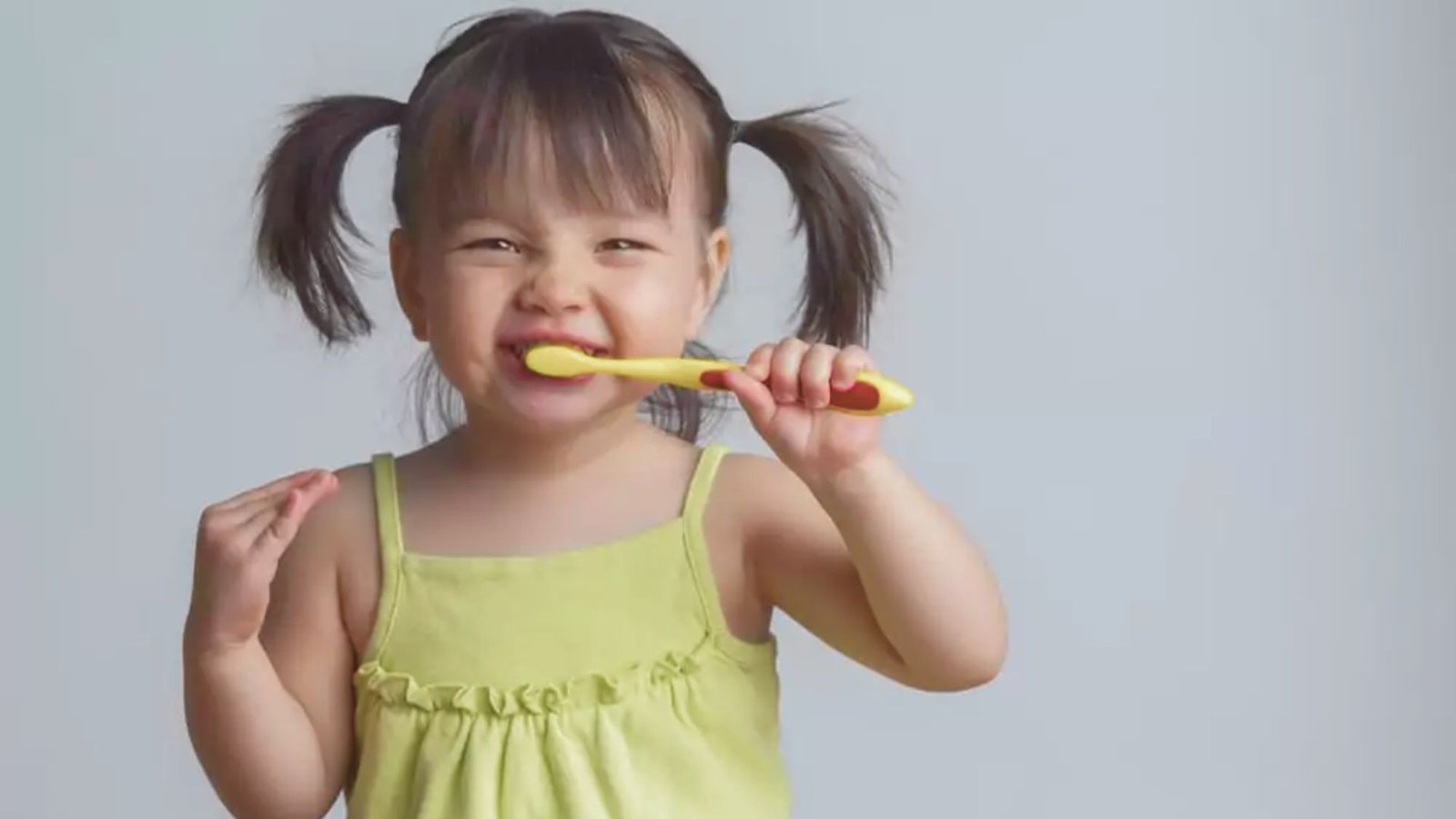 A young girl brushes her teeth with a bright yellow toothbrush, smiling in a bathroom setting.