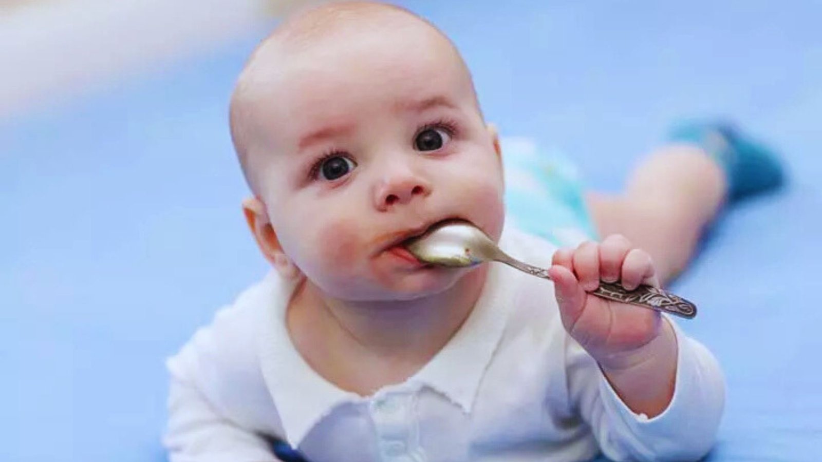 A baby lying on a blue blanket, playfully holding a spoon in its mouth, learning textures and tastes.