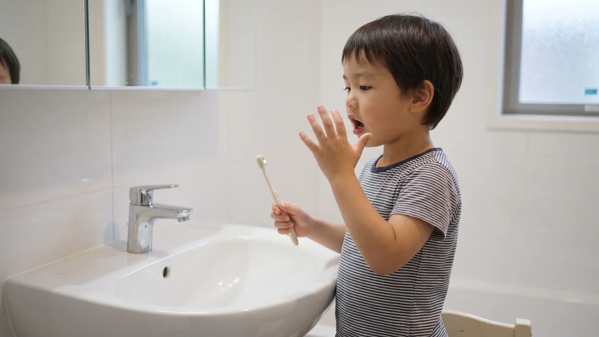 Young child holding toothbrush checking mouth in bathroom sink