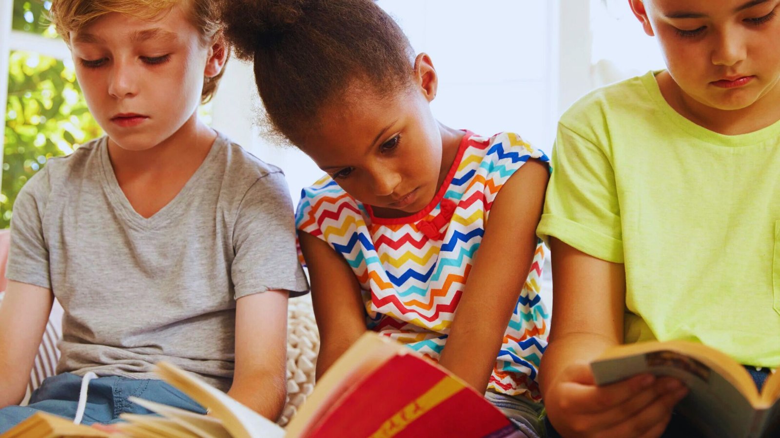 Three children sit on a couch, each engrossed in their own book, surrounded by a cozy living room setting.