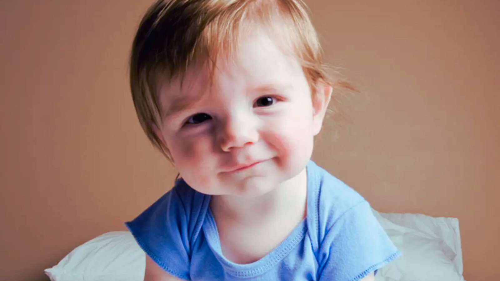 A baby wearing a blue shirt sits on a bed, looking curiously.