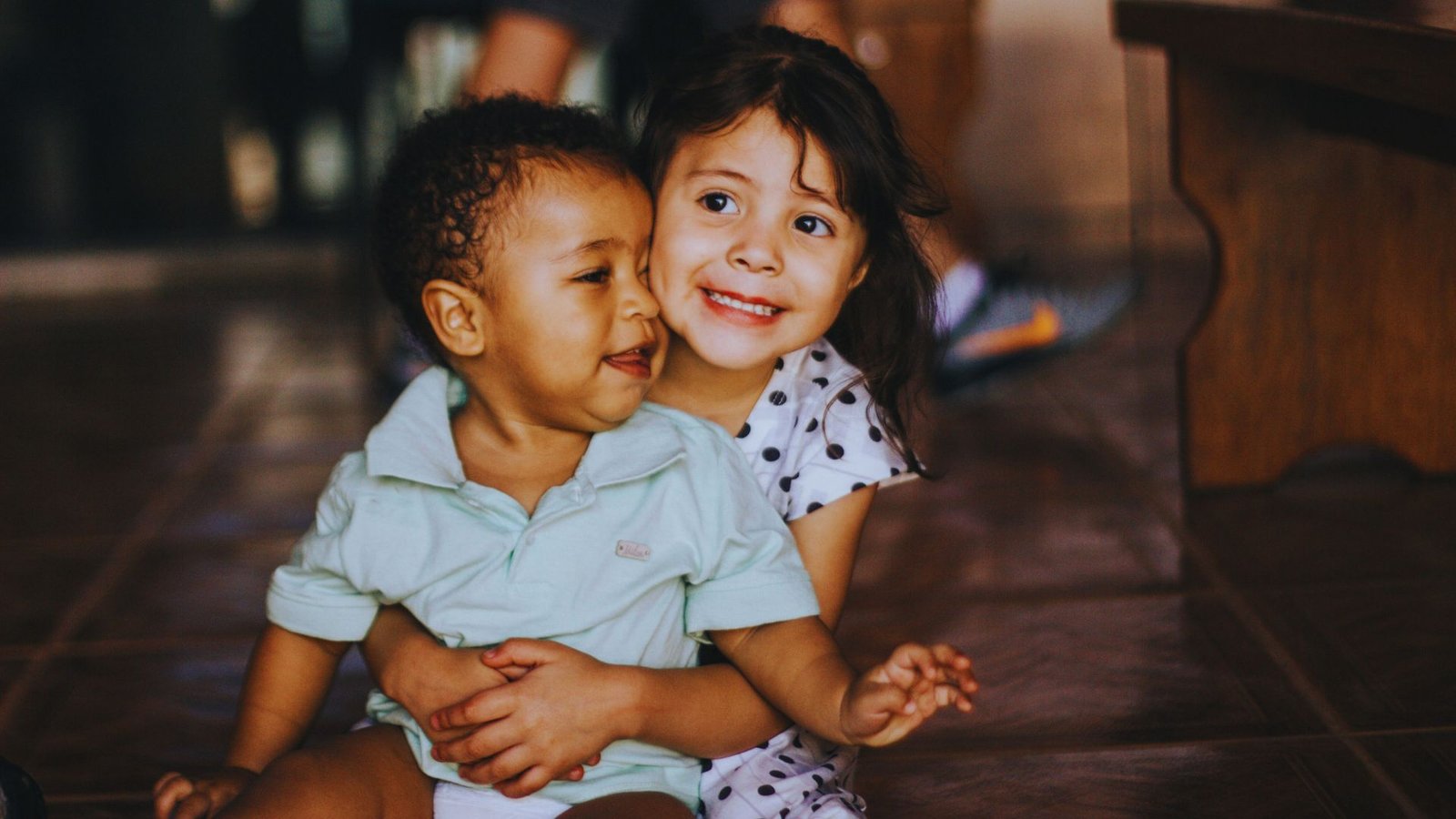 Two children sitting on the floor, one gently holding the other in a caring embrace.