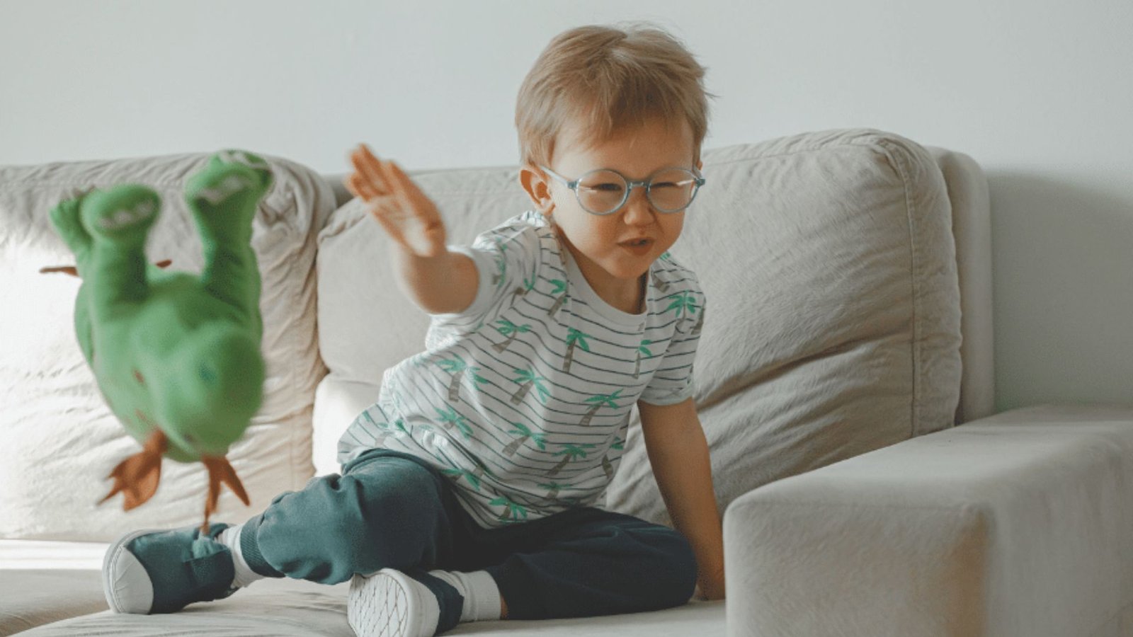 A young boy with glasses sits on a couch, holding a green toy and smiling happily.
