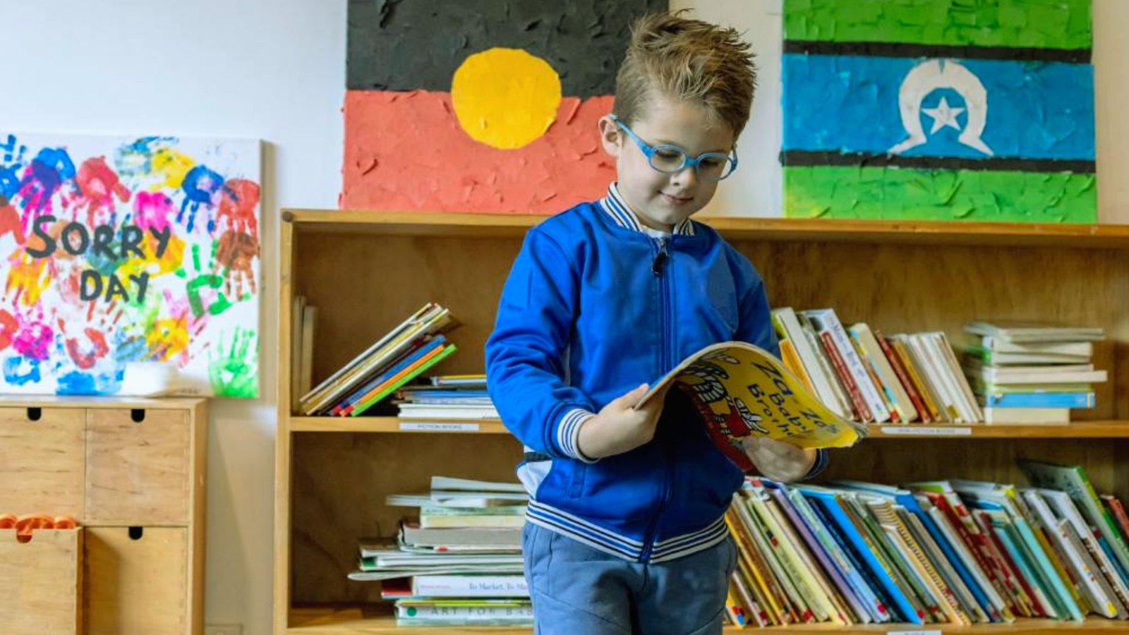 A young boy reads a book in a library, focused and surrounded by tall shelves of books.