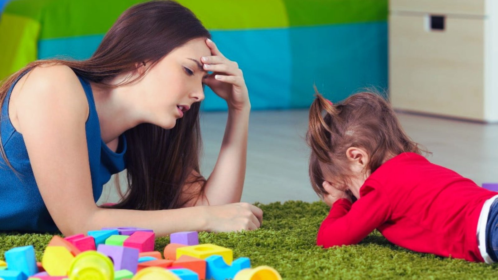 A woman and a child relax on the floor, various toys scattered around them in a cozy setting.