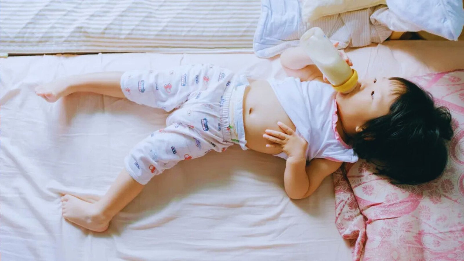 A baby lying on a bed, peacefully holding a bottle, surrounded by soft bedding.
