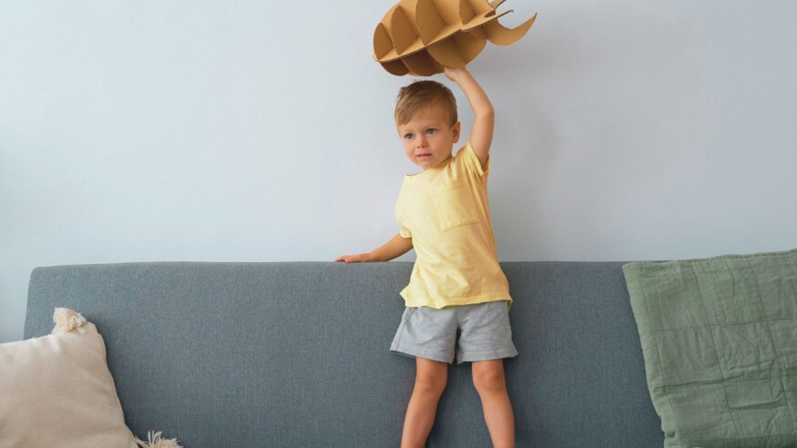 A little boy stands on a couch, holding a paper hat while throwing toys around him.
