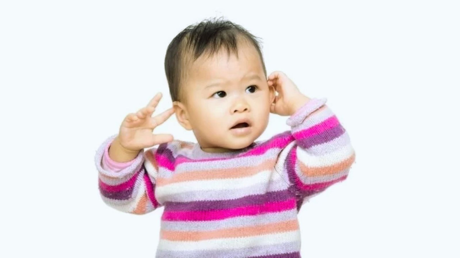 A baby stands with her hands on her ears, appearing startled or overwhelmed.