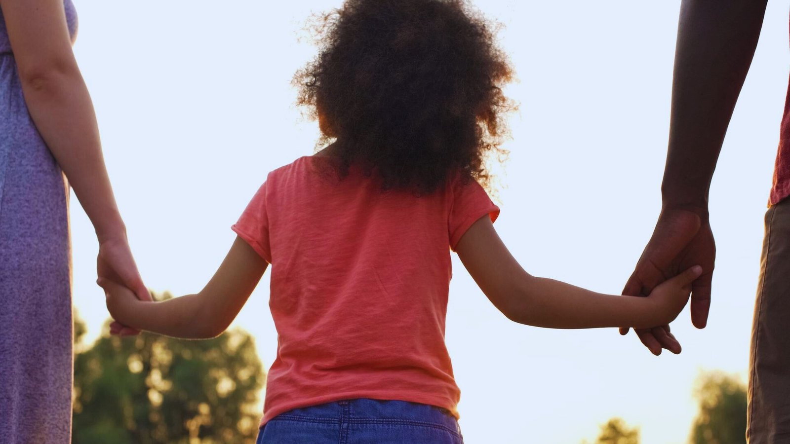 A young girl joyfully holds hands with two adults, symbolizing connection and support in a warm outdoor setting.