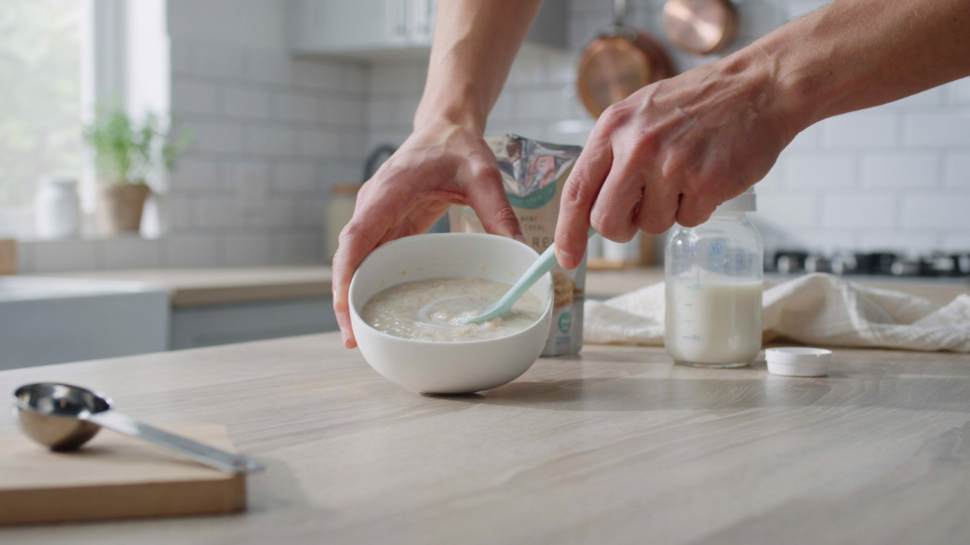 Parent preparing smooth rice cereal with baby feeding supplies in a kitchen.