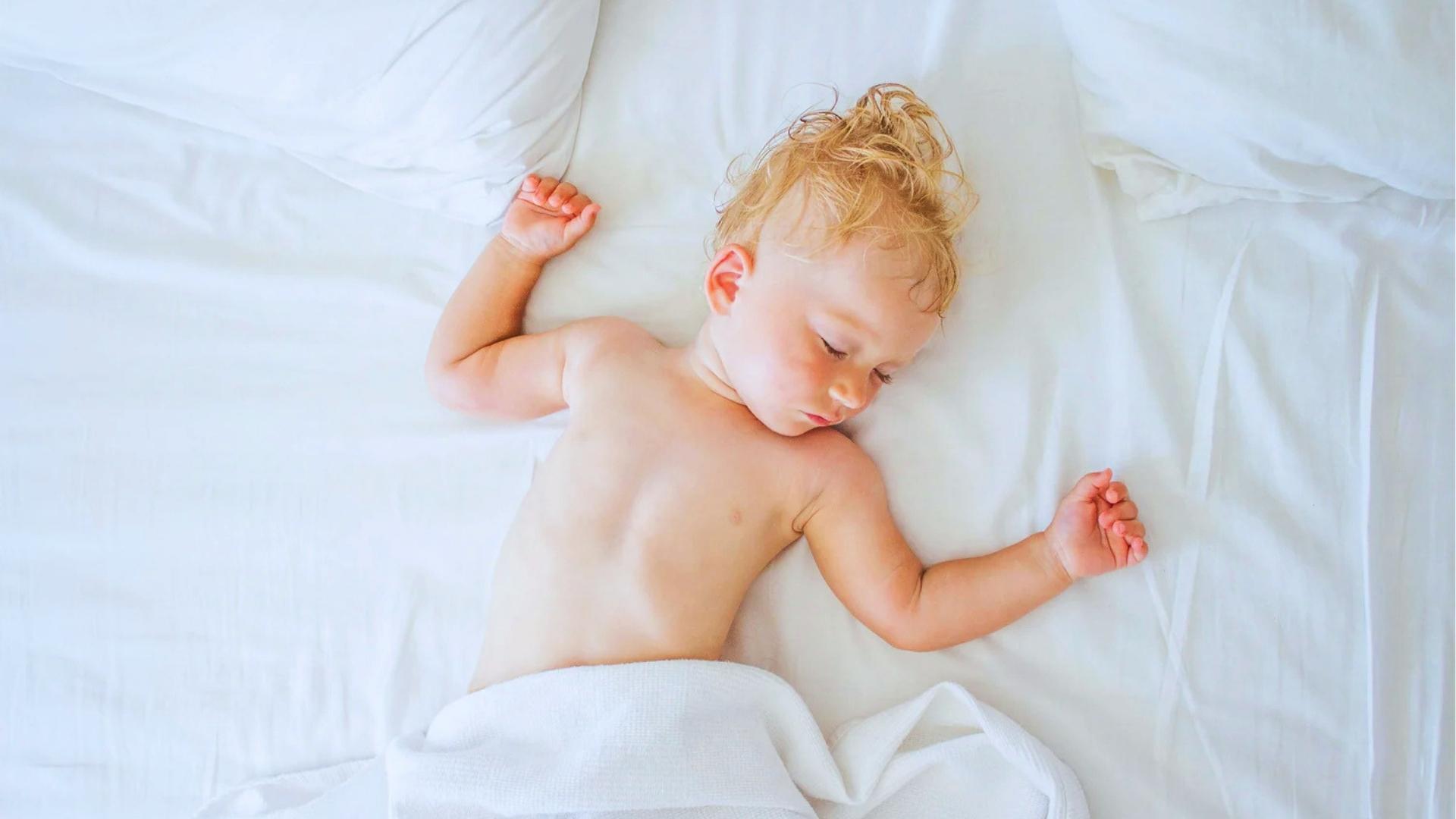Toddler with curly blonde hair sleeps on white sheets, face slightly flushed and damp.