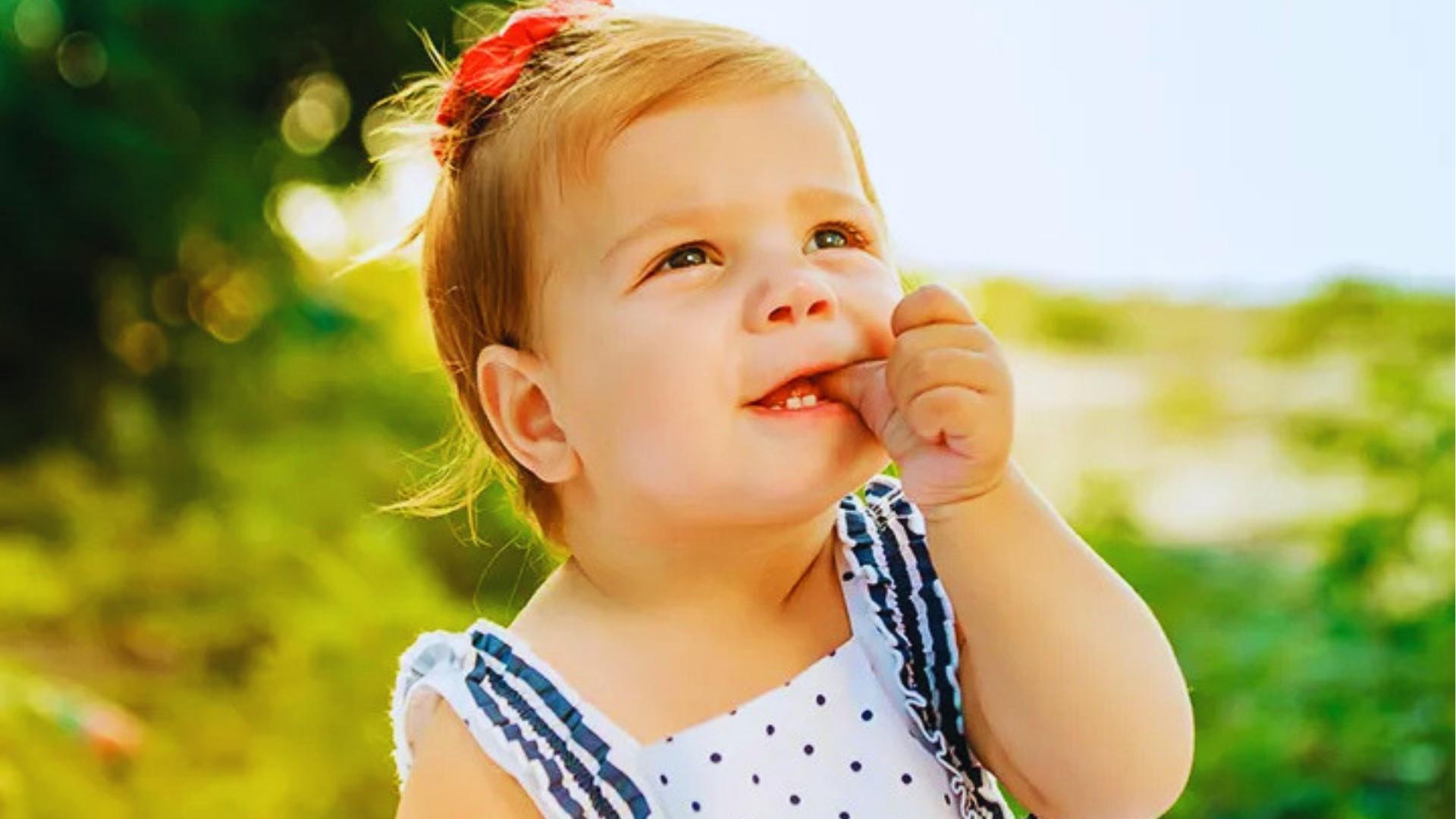 Smiling toddler in polka-dot dress with red bow sucks thumb outdoors in sunny park.