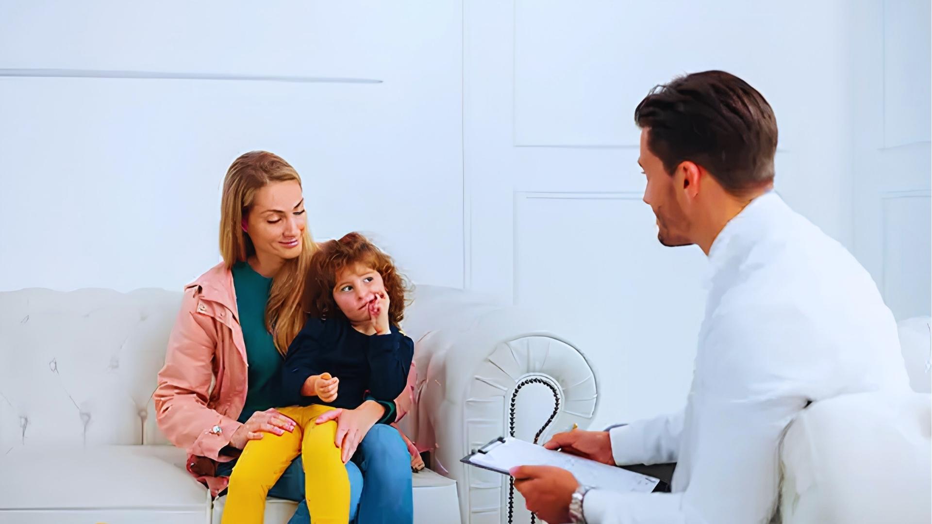 Smiling mother in pink jacket holds curly-haired toddler sucking thumb during consultation with male doctor.