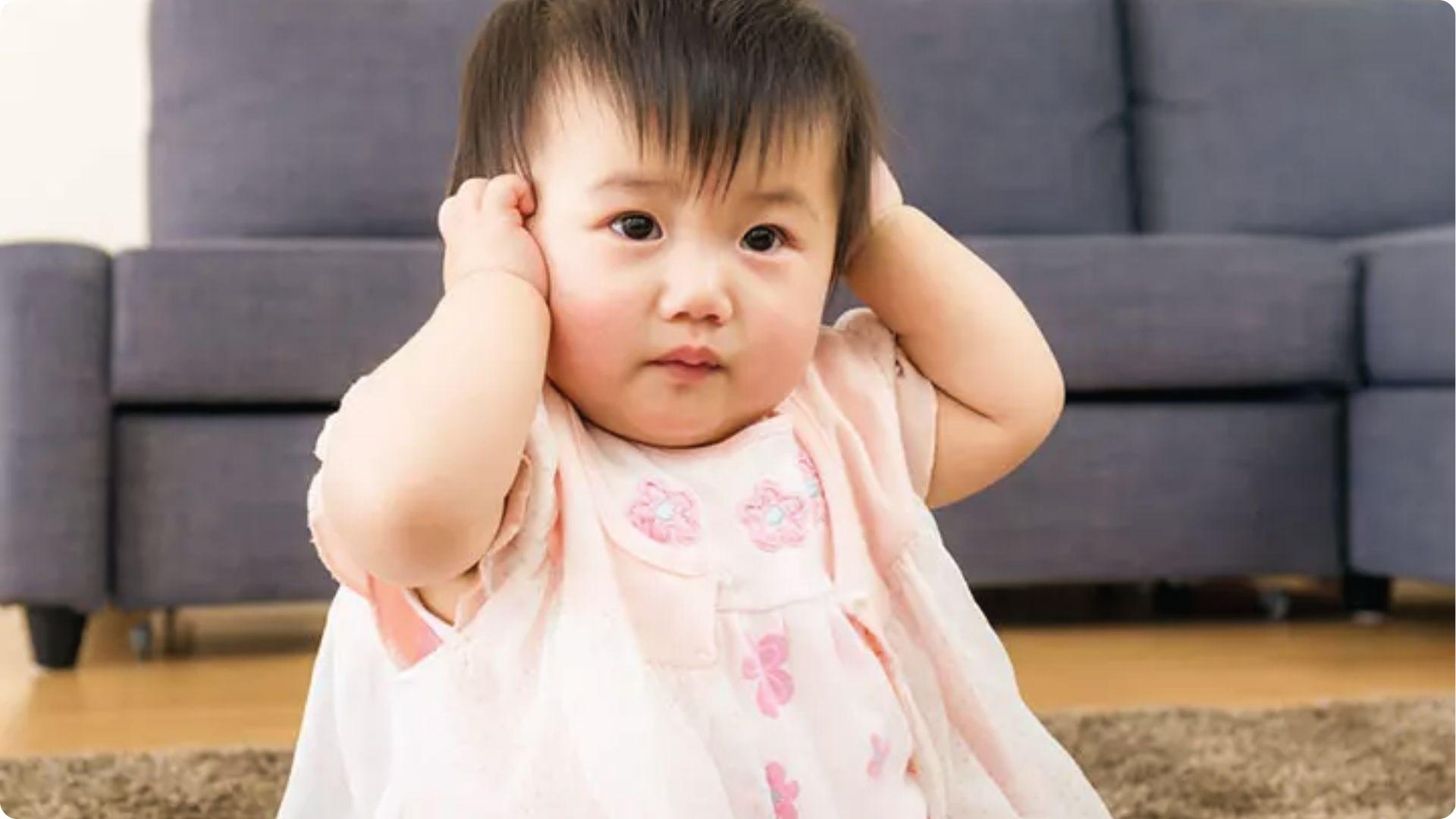 Cute baby in pink dress holding her ears and shaking her head.