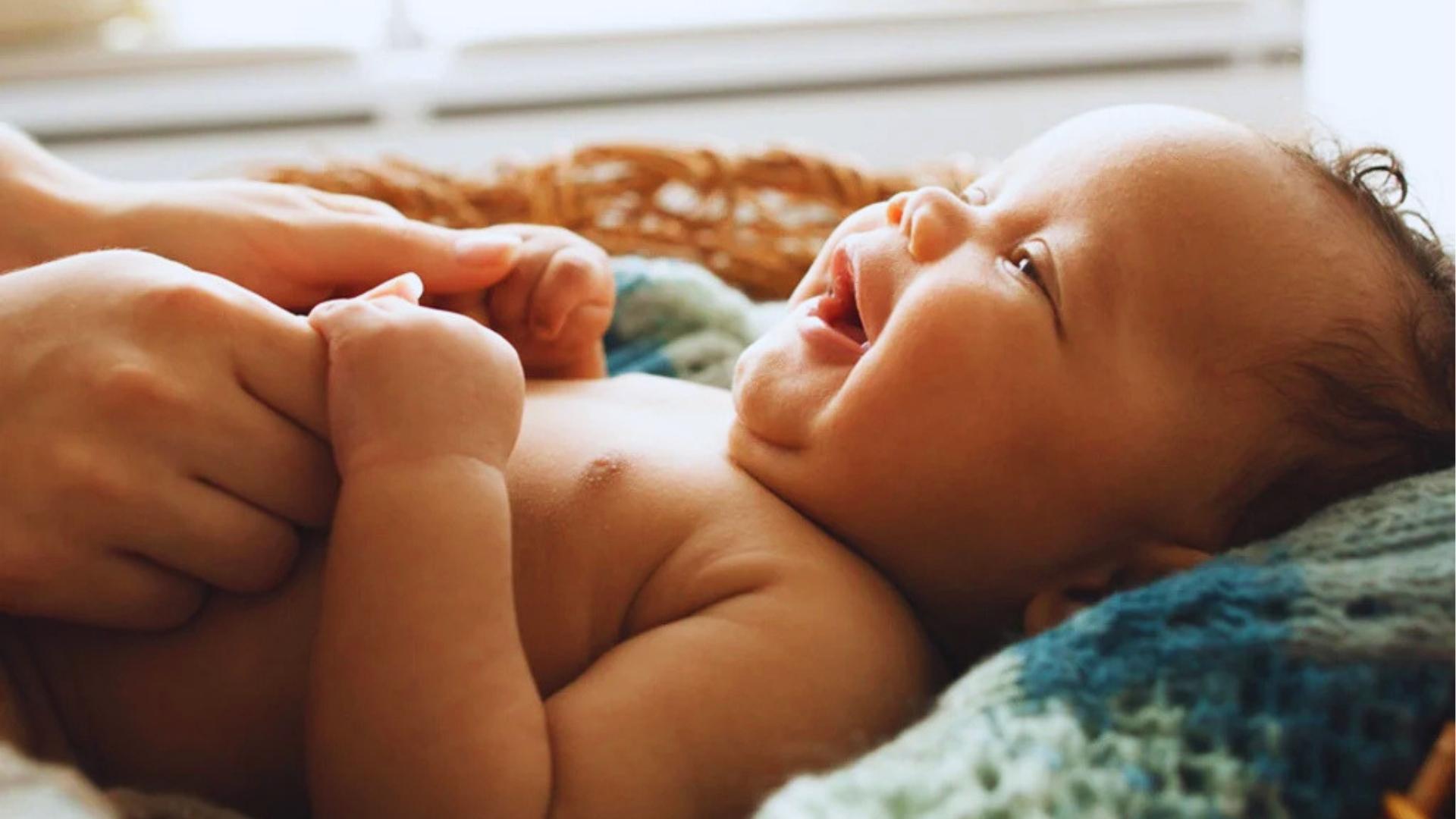 Smiling baby lies in basket, held by adult hands, looking up at caregiver happily.