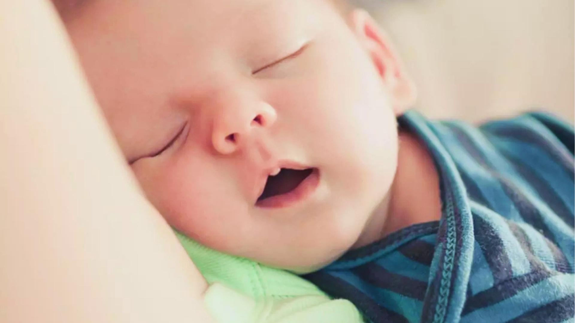 Close-up of sleeping baby in blue striped shirt and green bib, mouth open, eyes closed peacefully.