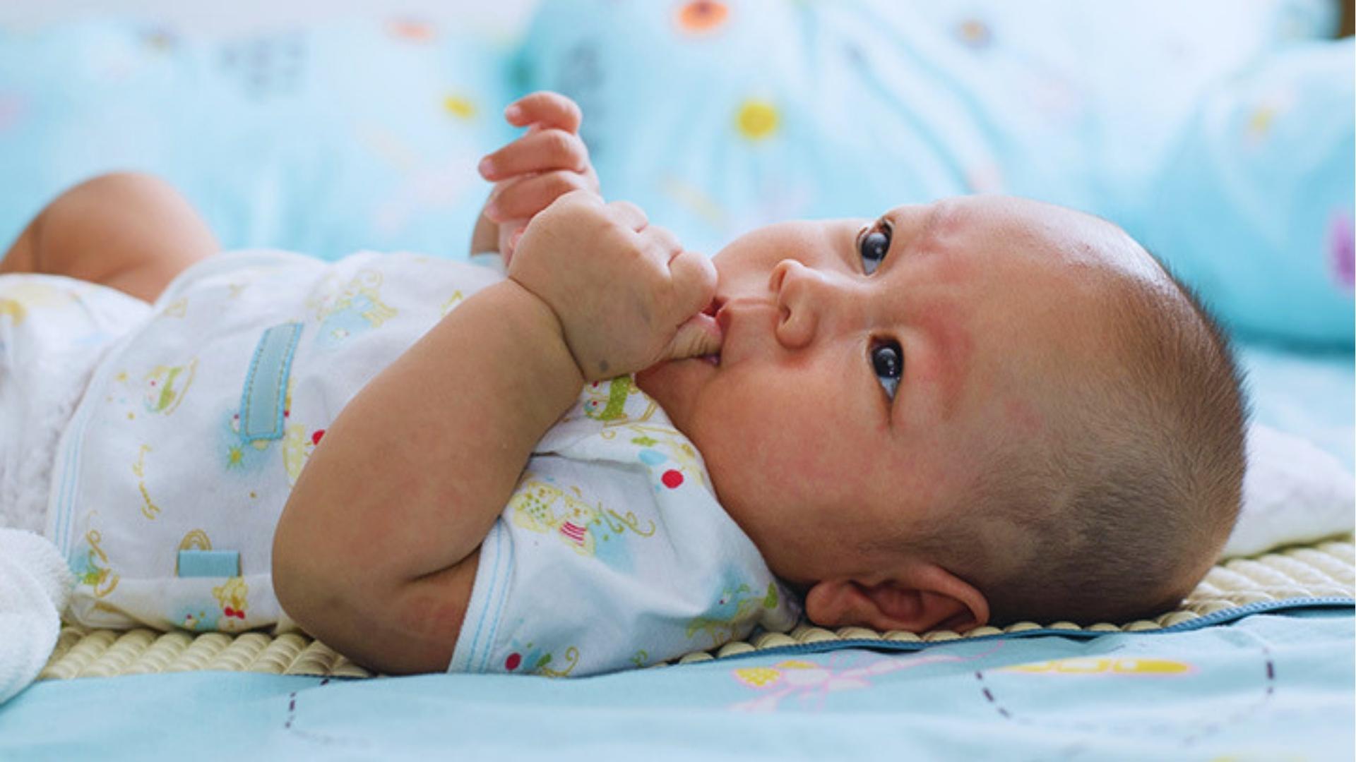 Baby in white patterned onesie lies on blanket sucking thumb, cheeks slightly flushed pink.