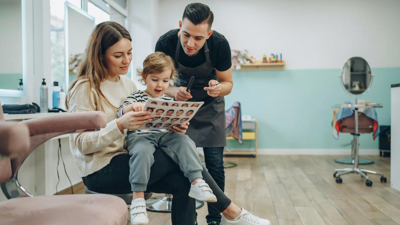 Mother helping choose the best hairstyle for her 2-year-old son at a kids salon.