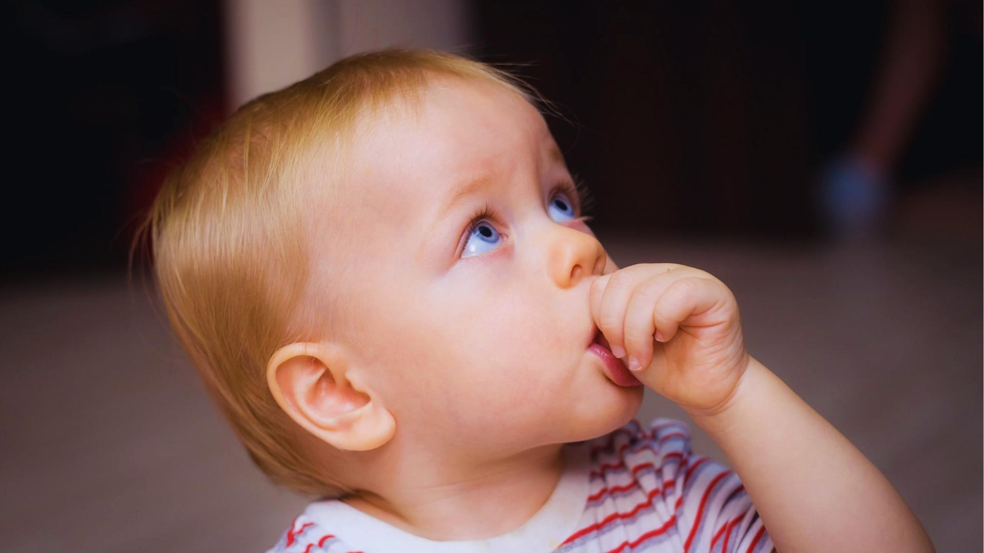 Blonde baby in striped shirt sucks thumb while gazing upward with bright blue eyes.