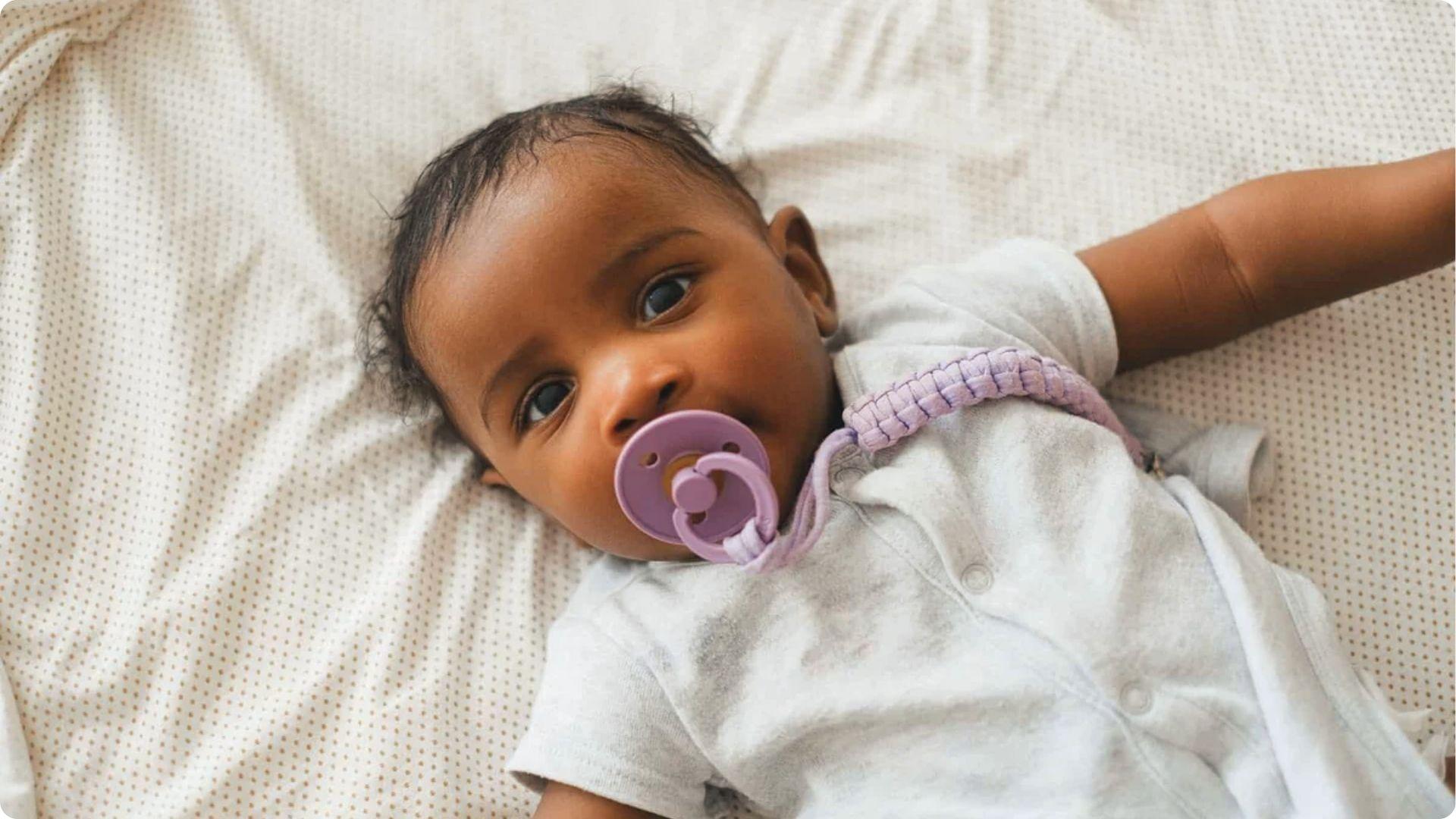 The baby lies on bed with a purple pacifier and clip, highlighting safe pacifier use for infant comfort.