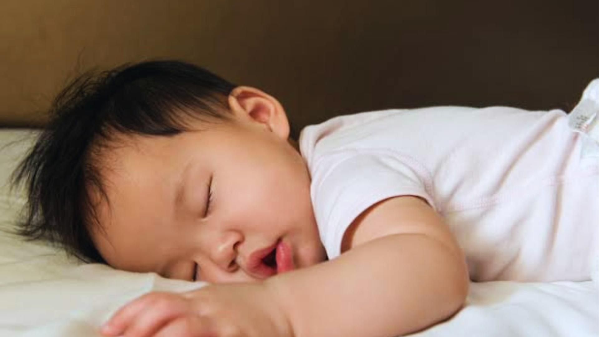 Baby with dark hair sleeps on stomach wearing white onesie, mouth slightly open on bed.