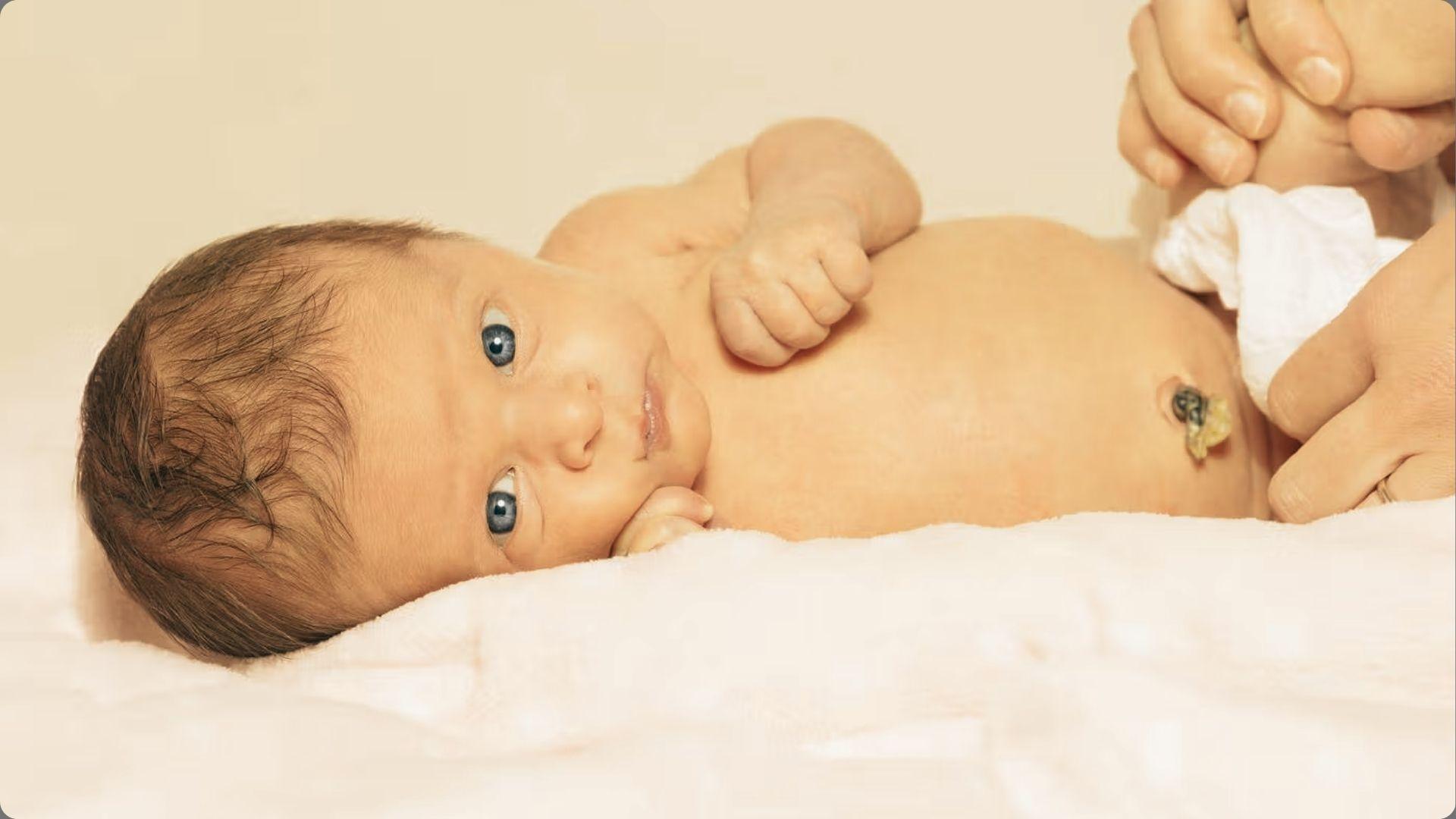 Newborn with blue eyes lying on blanket, dried umbilical cord stump visible on belly, adult hands nearby