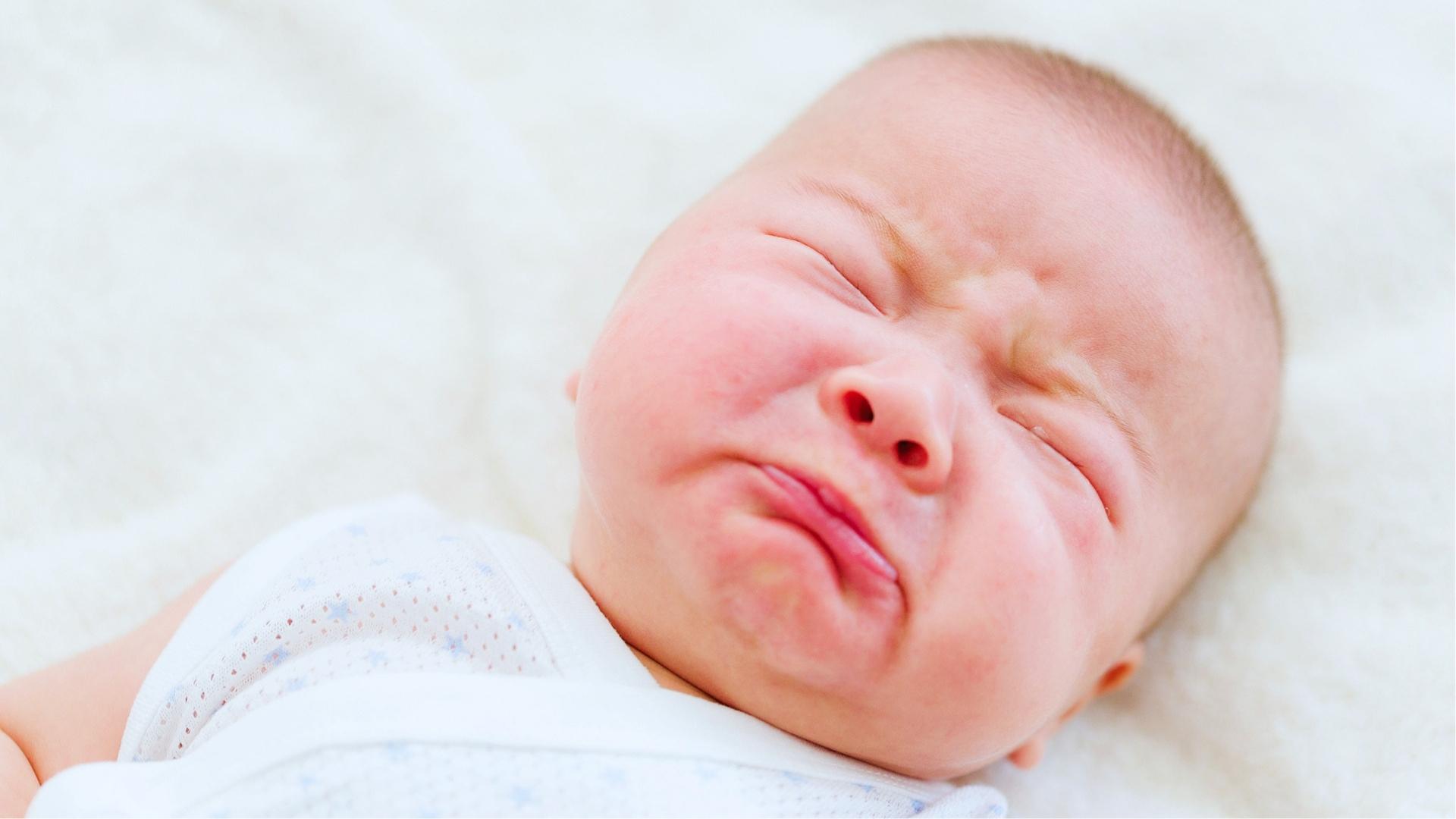 Baby in white onesie scrunches face while crying, cheeks flushed red on white blanket.