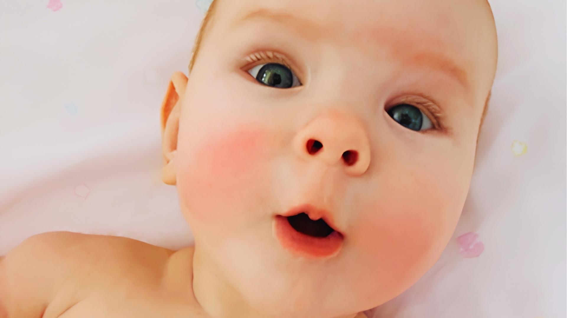 Baby with blue eyes and pink rosy cheeks lies on white blanket, mouth slightly open.