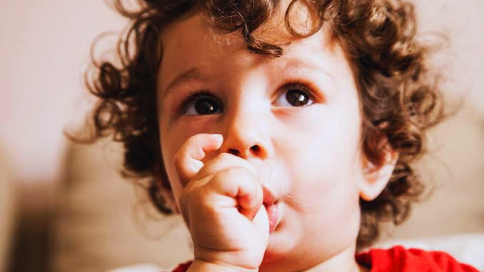Close-up of curly-haired toddler in red shirt sucking thumb, looking upward with big eyes.