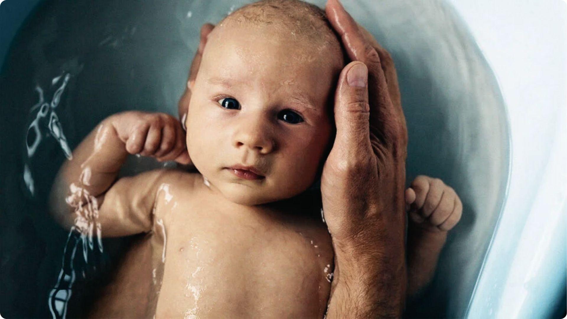 Baby in a bath with adult hand supporting head; head shaking can be reaction to water or ear sensation.