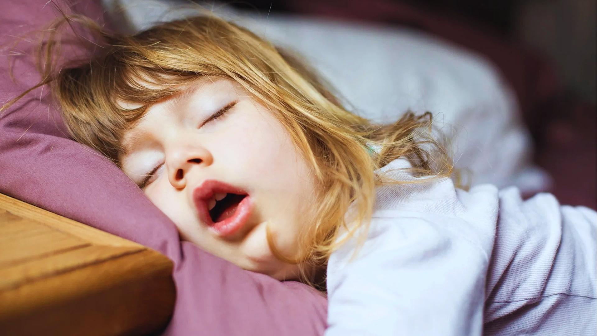 Young child with open mouth sleeps on side, head resting on purple pillow in bed.