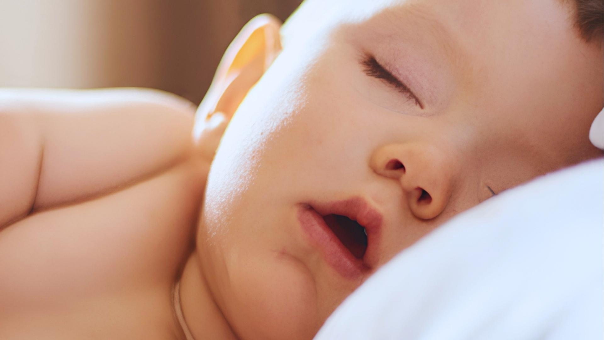 Close-up of sleeping baby with open mouth, soft sunlight on face, resting on white blanket.