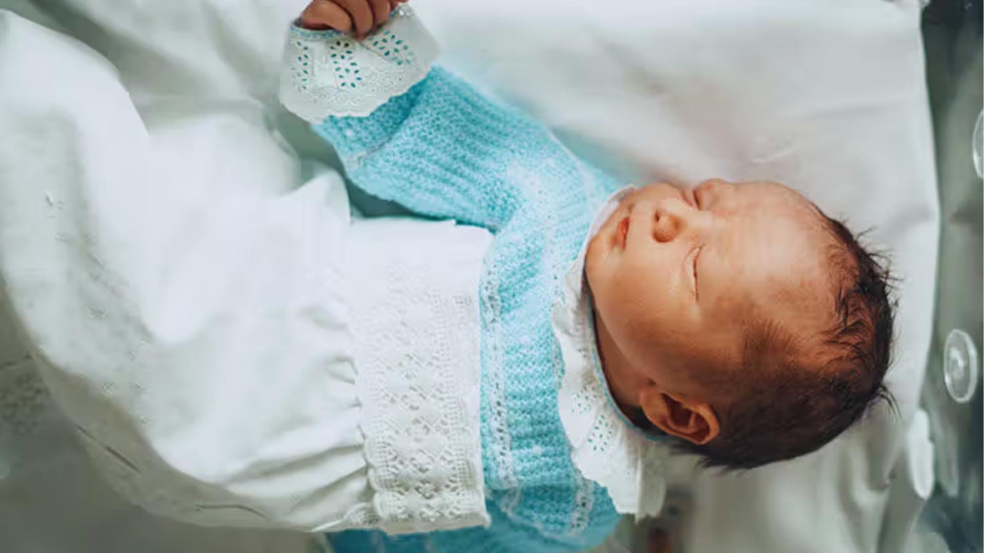 Newborn in blue knit sweater and white lace dress sleeps peacefully with red face.