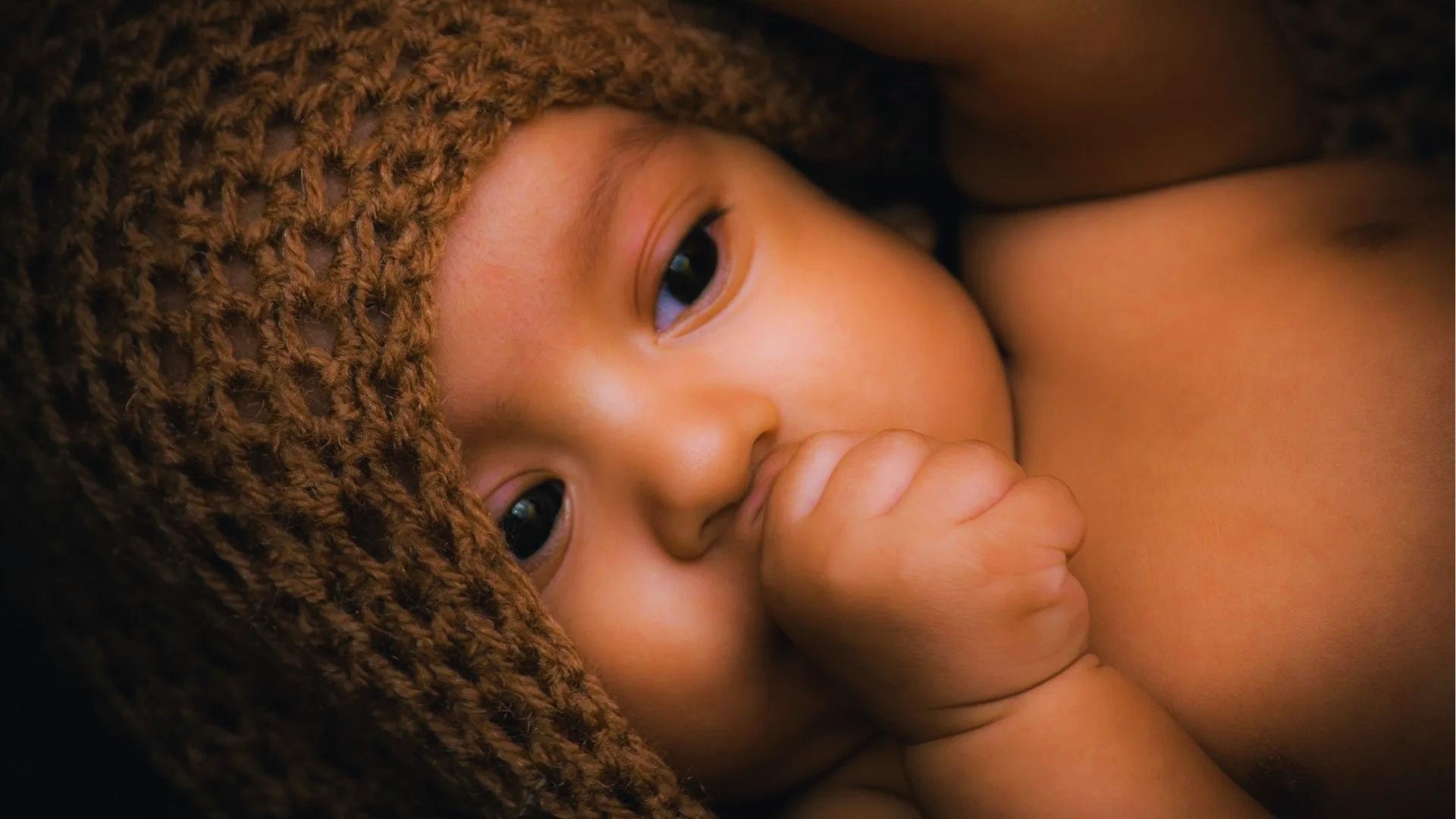 Newborn in brown knit hat sucks thumb while wrapped in matching crocheted blanket, looking at camera.