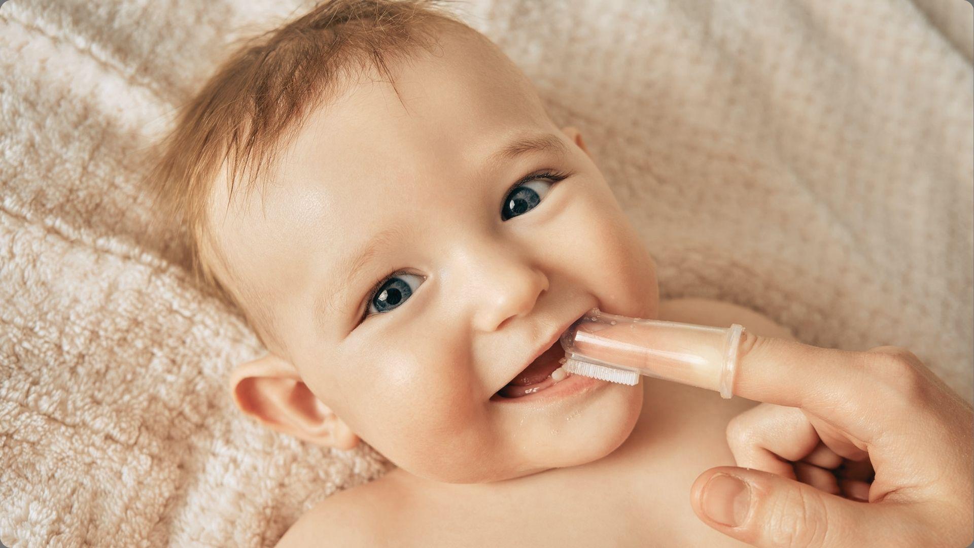 Smiling blue-eyed baby with light brown hair lying on blanket while adult brushes teeth with silicone finger toothbrush