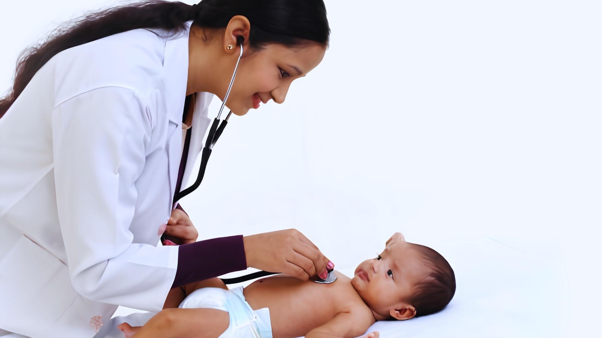 Smiling female doctor in white coat uses stethoscope on baby lying on exam table.
