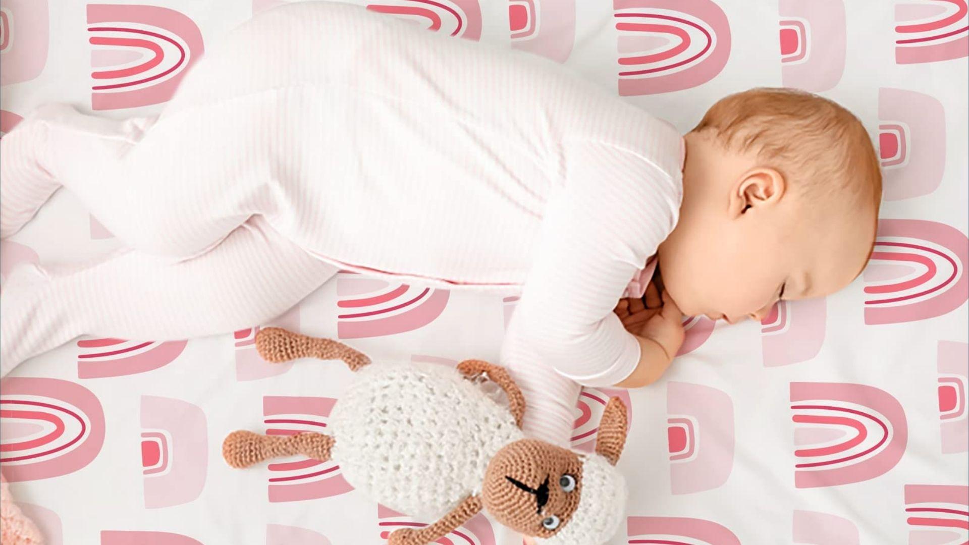 Baby in white onesie sleeping in bassinet on pink rainbow sheet with crocheted sheep toy
