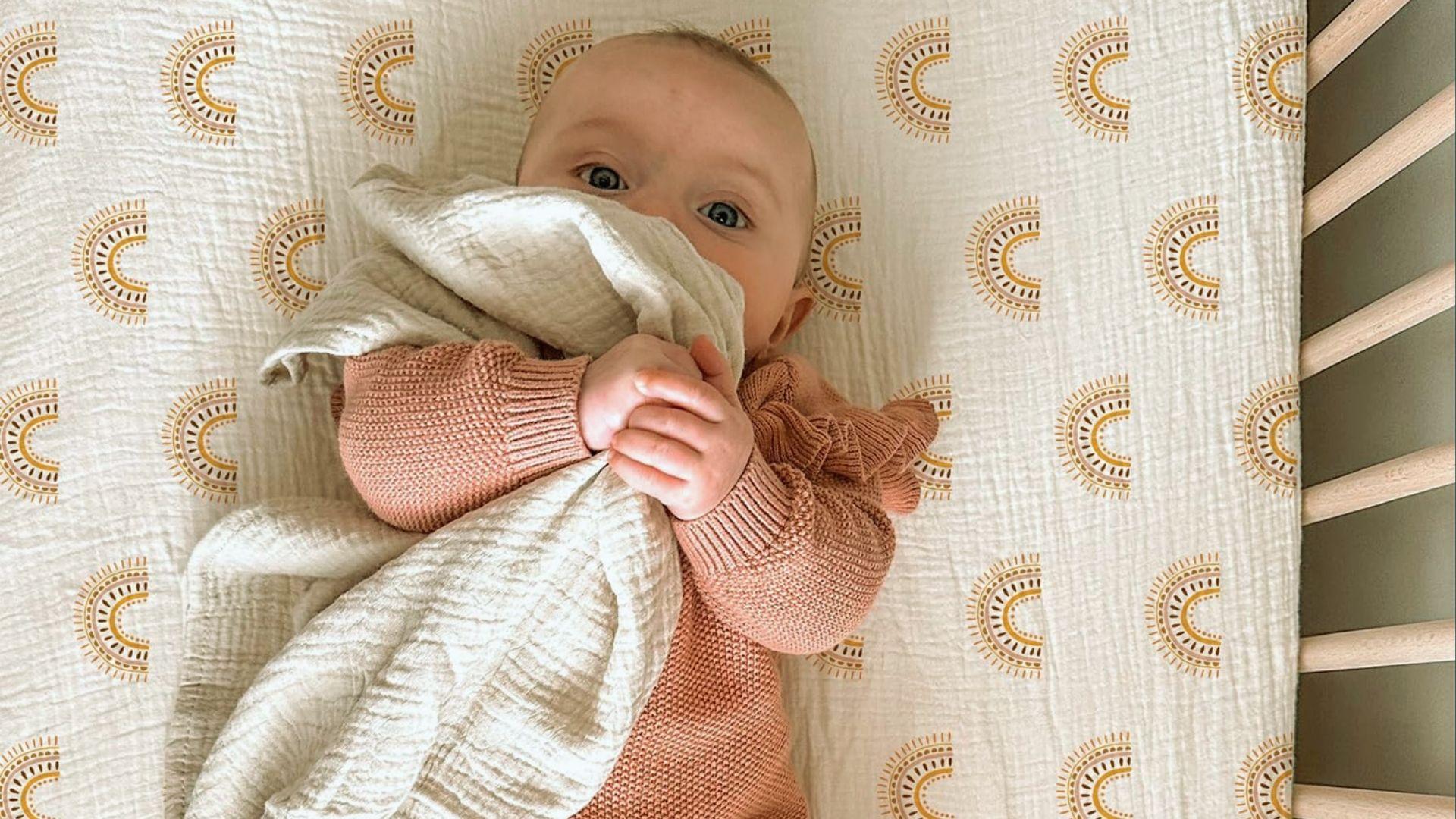 Baby in peach knit sweater holding cloth over mouth lying in bassinet with sun-pattern sheet