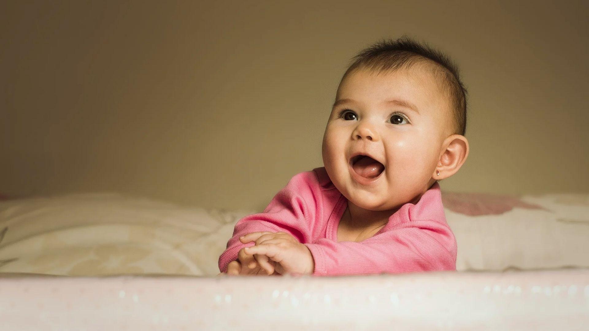 Happy baby girl in pink onesie lying on bed smiling joyfully during tummy time