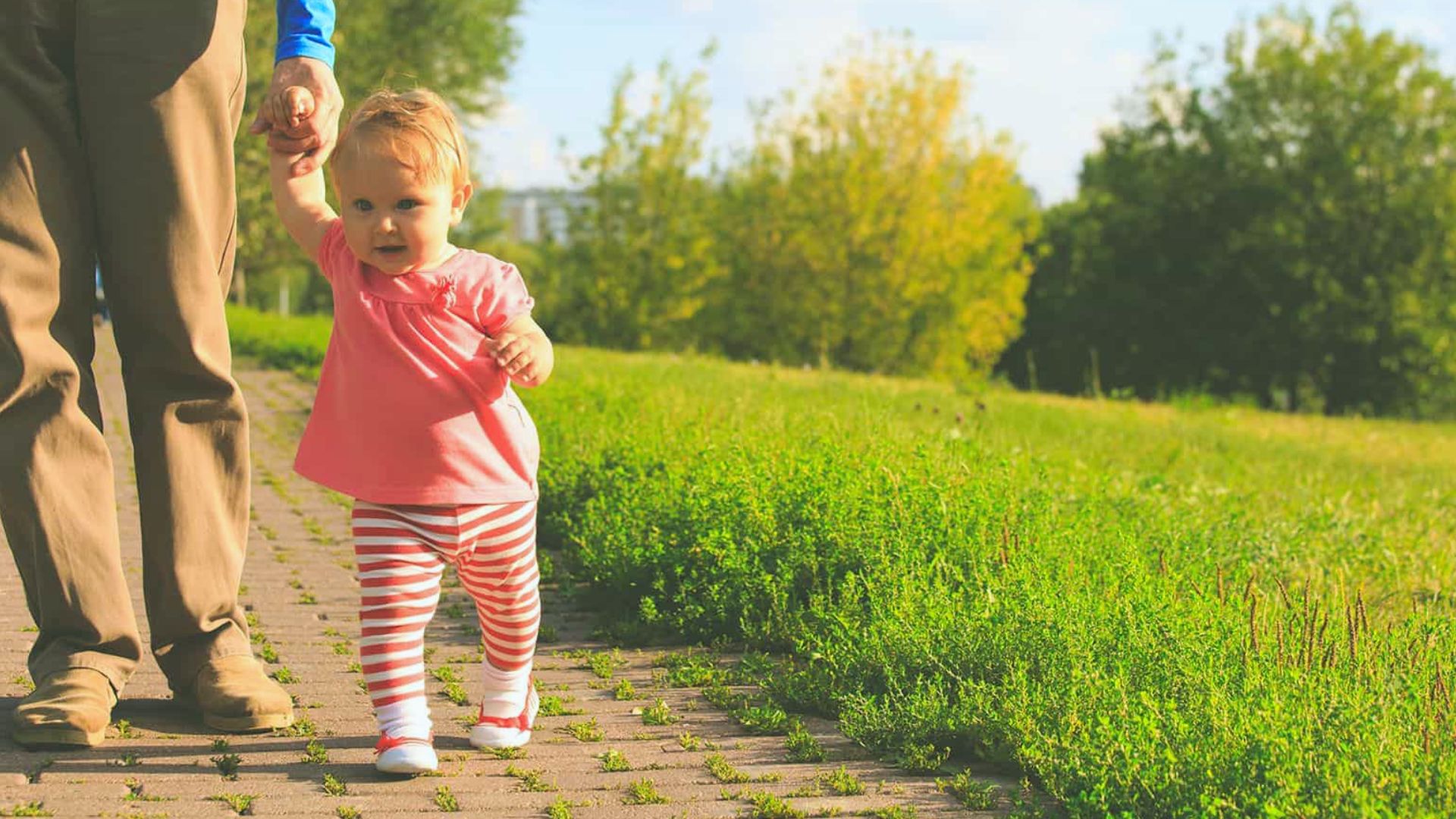 Toddler in pink shirt and striped leggings walking on path holding adult's hand