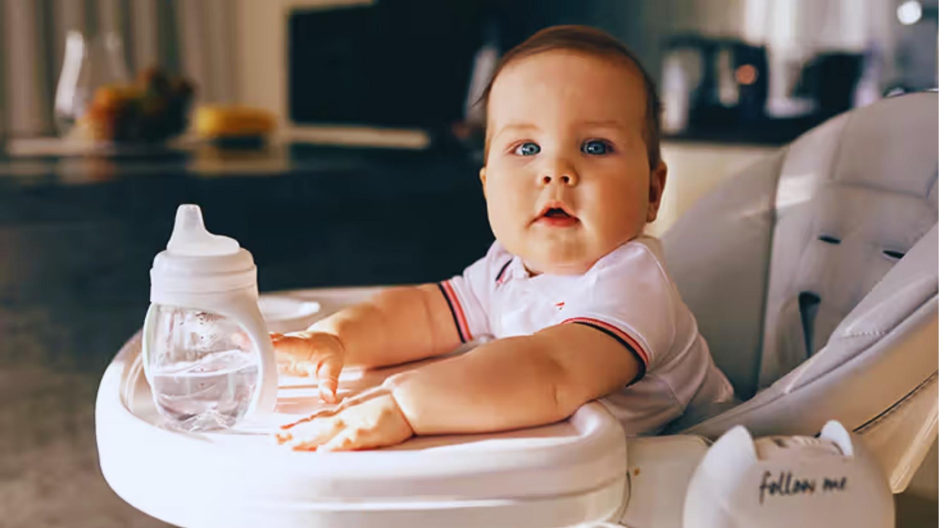 Cute baby sitting in high chair beside sippy bottle looking surprised during mealtime feeding today.