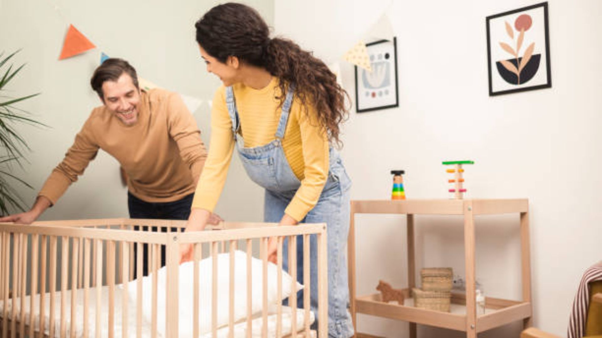 Happy couple smiling while assembling light wooden baby crib together in bright nursery with toys and framed art on wall.