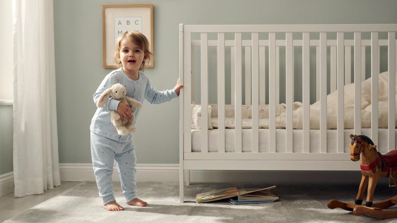 A toddler in blue pajamas holding a stuffed rabbit standing beside a white crib in a bright nursery after waking up from a nap.