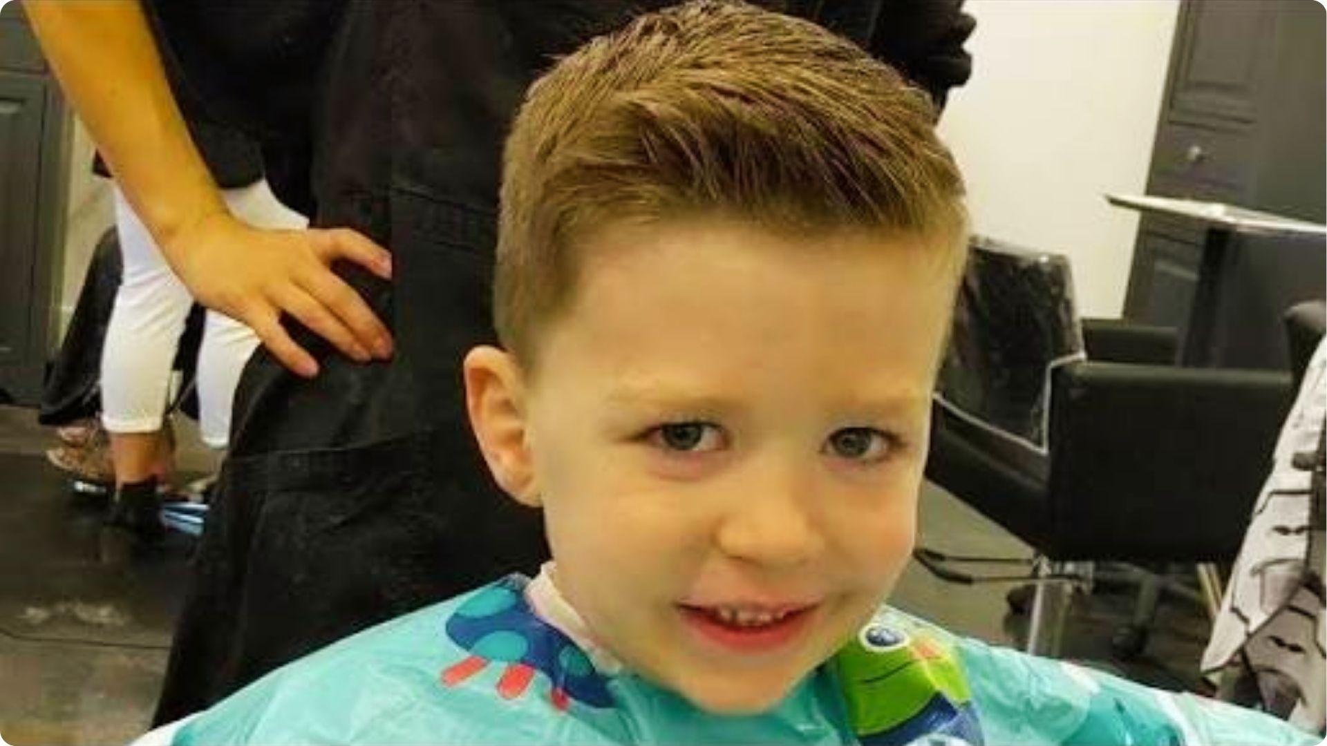 Toddler in barber chair with neat short side-part comb-over, fresh trim, classic baby boy haircut.